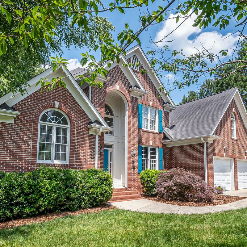 A red brick two-story house behind shrubs and a green lawn